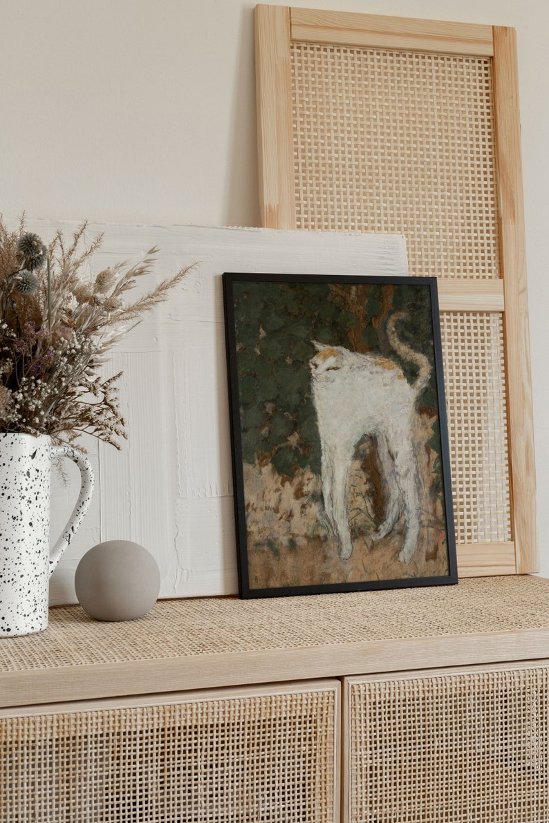 May include: A framed painting of a white cat with a brown tail, standing in a green and brown landscape. The painting is hanging on a wall with a wicker panel behind it. A vase with dried flowers and a round, gray object are on a wooden shelf below the painting.