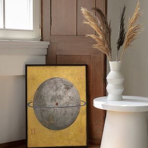 Op de afbeelding: Een ingelijste print van een hemelbol met een gouden achtergrond en een blauwe ring rond de bol. De bol is grijs en heeft een klein rood stipje in het midden. De print is zwart ingelijst en hangt aan een muur.
