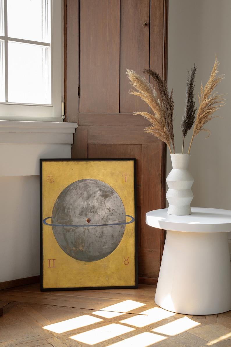 Op de afbeelding: Een ingelijste print van een hemelbol met een gouden achtergrond en een blauwe ring rond de bol. De bol is grijs en heeft een klein rood stipje in het midden. De print is zwart ingelijst en hangt aan een muur.