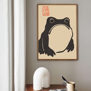 May include: A black and white illustration of a frog with a red stamp in the top left corner. The stamp has Japanese characters on it. The illustration is on a beige background.