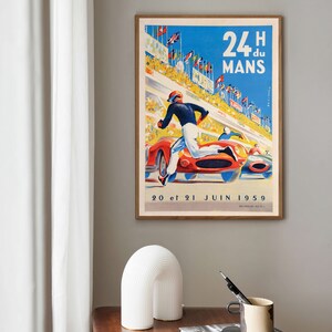 May include: Vintage poster advertising the 24 Hours of Le Mans race, featuring a red race car and a driver running on the track. The poster is framed and has a blue background with white text that reads "24H du MANS" and "20 et 21 JUIN 1959".