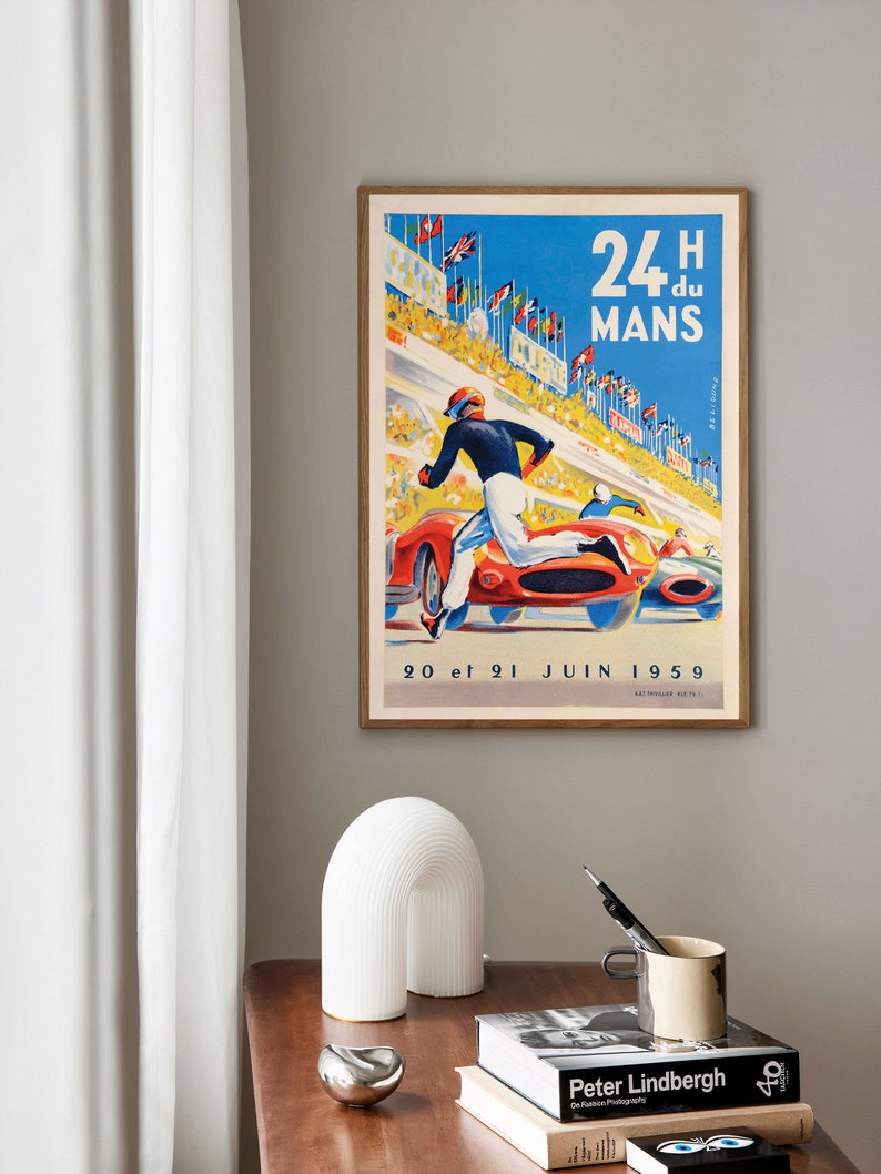 May include: Vintage poster advertising the 24 Hours of Le Mans race, featuring a red race car and a driver running on the track. The poster is framed and has a blue background with white text that reads "24H du MANS" and "20 et 21 JUIN 1959".