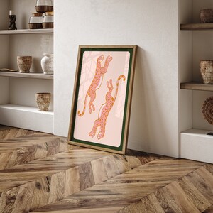 May include: A framed print of two pink leopards with orange spots leaping in the air against a light pink background. The frame is brown with a dark green mat.