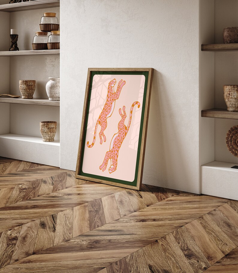 May include: A framed print of two pink leopards with orange spots leaping in the air against a light pink background. The frame is brown with a dark green mat.