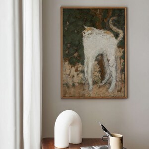 May include: A white cat painting in a wooden frame hangs on a light beige wall above a wooden table with a white ceramic lamp, a silver metal object, and a stack of books. The top book has the title "Peter Lindbergh" on the cover.