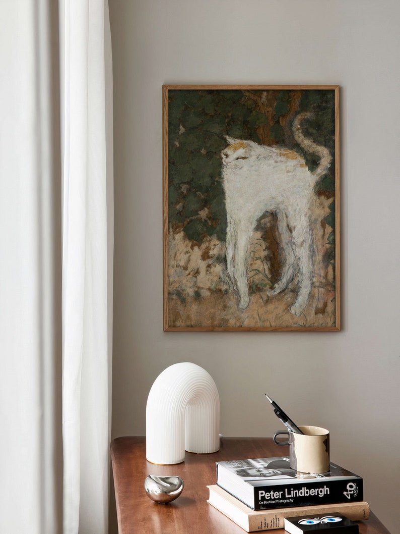 May include: A white cat painting in a wooden frame hangs on a light beige wall above a wooden table with a white ceramic lamp, a silver metal object, and a stack of books. The top book has the title "Peter Lindbergh" on the cover.