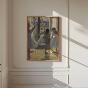 May include: A framed print of a painting by Edgar Degas featuring three ballerinas in white tutus. The painting is set in a dance studio with a window in the background. The text "DEGAS" is printed in white at the bottom of the print.