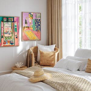 May include: A white bed with a beige knitted throw blanket and a straw hat. The bed is styled with white pillows and a beige pillow with a woven texture. Two framed paintings of colorful abstract art hang on the wall behind the bed.