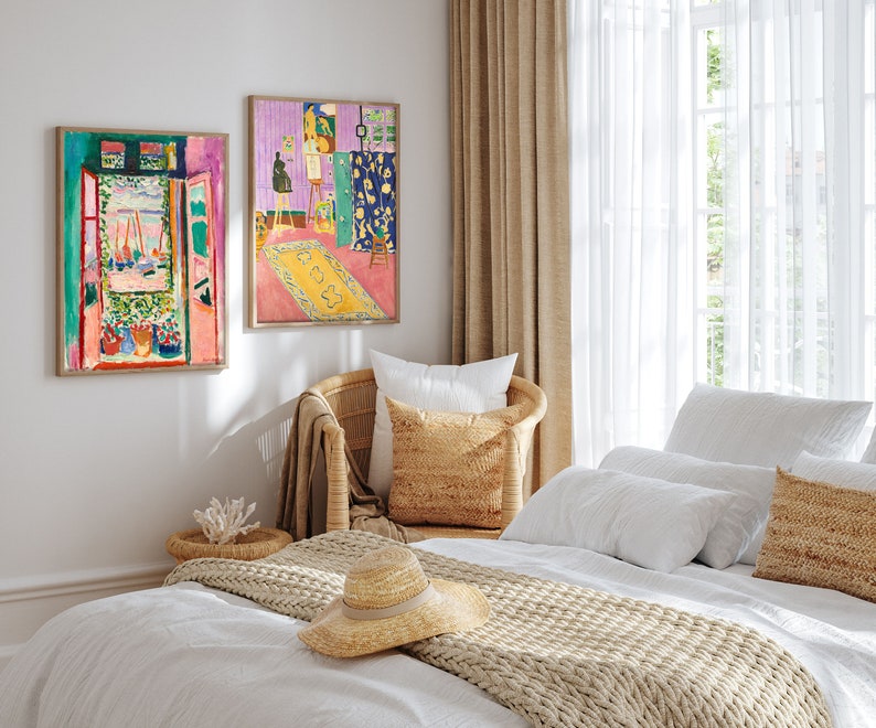 May include: A white bed with a beige knitted throw blanket and a straw hat. The bed is styled with white pillows and a beige pillow with a woven texture. Two framed paintings of colorful abstract art hang on the wall behind the bed.