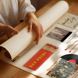 May include: A person is unrolling a large, white poster on a wooden surface. A red matchbox with the text "BAR PRIMI" in yellow is visible, along with a vinyl record and two matches. The scene suggests a vintage or retro theme.