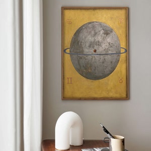 Op de afbeelding: Een ingelijste print van een hemelbol met een blauwe ring eromheen, tegen een gele achtergrond. De bol is grijs en heeft een rode stip in het midden. De print is omgeven door een houten lijst.