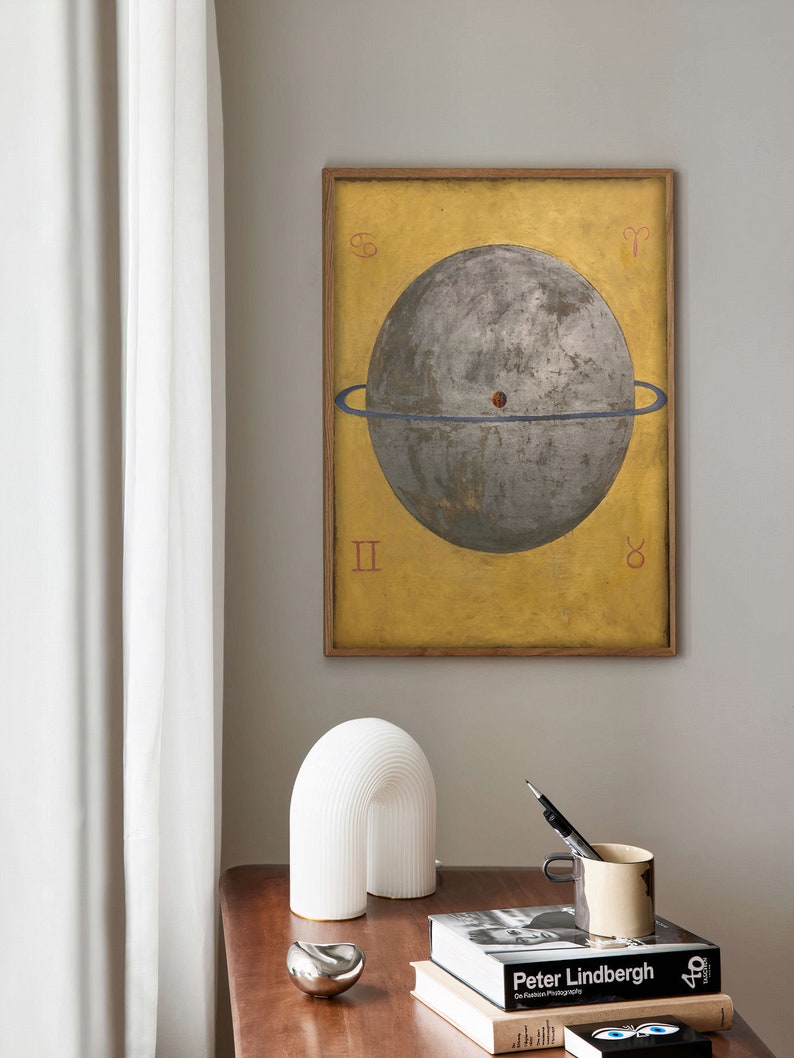 Op de afbeelding: Een ingelijste print van een hemelbol met een blauwe ring eromheen, tegen een gele achtergrond. De bol is grijs en heeft een rode stip in het midden. De print is omgeven door een houten lijst.