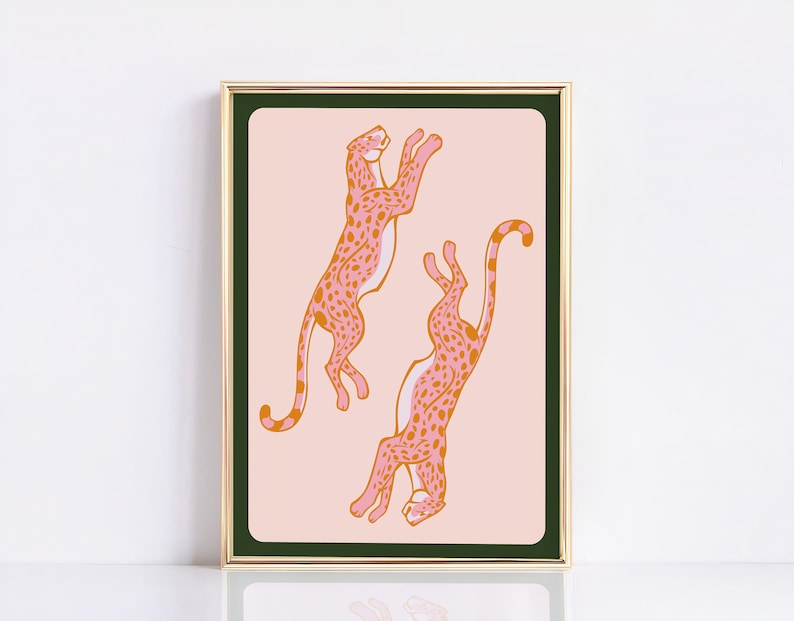 May include: A framed print with a pink background featuring two pink and orange cheetahs with black spots. The cheetahs are in a playful pose, one leaping and the other with its tail curled up.