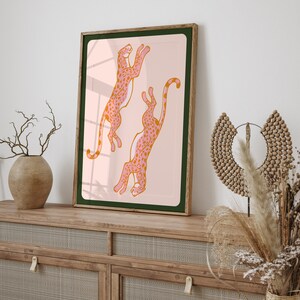 May include: A framed print of two pink and orange leopards leaping in the air against a light pink background. The frame is brown wood with a dark green mat.