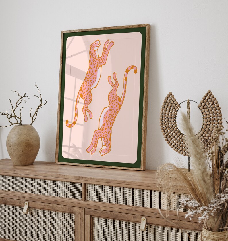 May include: A framed print of two pink and orange leopards leaping in the air against a light pink background. The frame is brown wood with a dark green mat.