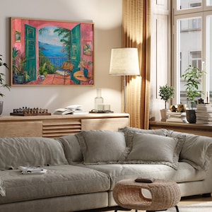 May include: A living room with a light grey couch, two pillows, and a small wicker table. A framed painting of a balcony overlooking a blue ocean is hanging on the wall. The painting is in a pink frame.