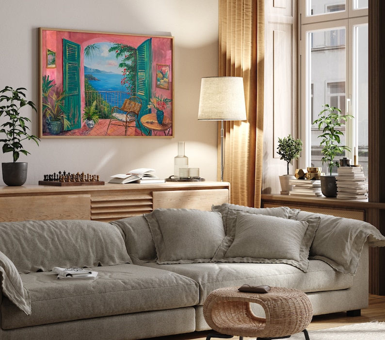 May include: A living room with a light grey couch, two pillows, and a small wicker table. A framed painting of a balcony overlooking a blue ocean is hanging on the wall. The painting is in a pink frame.