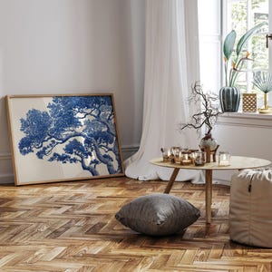 May include: A framed art print with a blue and white tree illustration. The artwork is in a light wooden frame, set against a white wall. The room features a wooden floor, a small round table, and a window with sheer curtains. The room is decorated with candles.