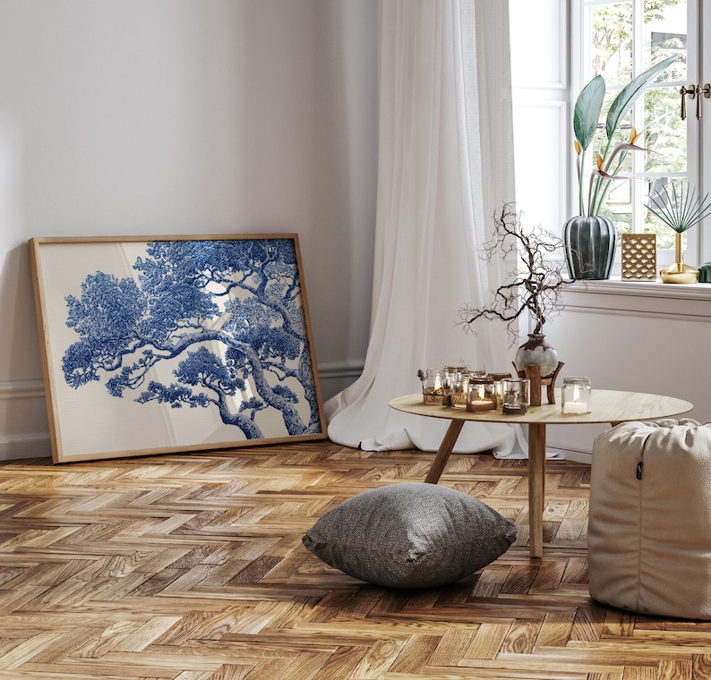 May include: A framed art print with a blue and white tree illustration. The artwork is in a light wooden frame, set against a white wall. The room features a wooden floor, a small round table, and a window with sheer curtains. The room is decorated with candles.