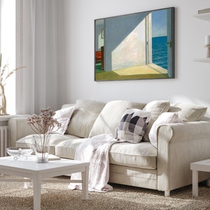 May include: A white sofa with a throw blanket and cushions in a living room setting. A framed painting of a seascape hangs on the wall above the sofa.