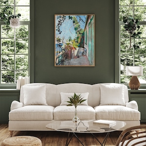 May include: A white linen sofa with three pillows sits in a living room with a green wall. A glass coffee table with a book on top sits in front of the sofa. A painting of a green and blue outdoor scene hangs on the wall behind the sofa.