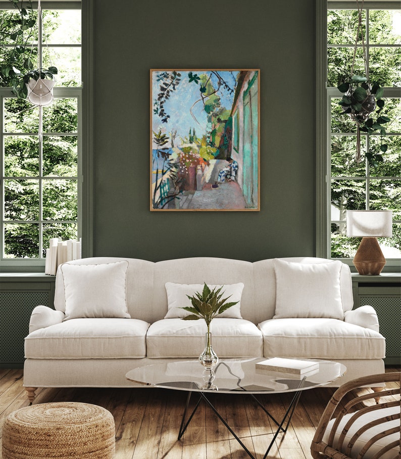 May include: A white linen sofa with three pillows sits in a living room with a green wall. A glass coffee table with a book on top sits in front of the sofa. A painting of a green and blue outdoor scene hangs on the wall behind the sofa.