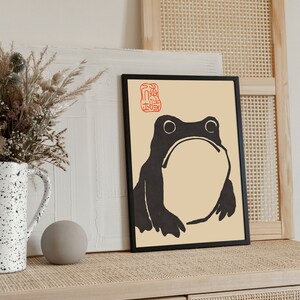 May include: Black and white illustration of a frog in a black frame on a light wood surface. The frog is on a cream background with a red stamp in the top left corner. The surface is a light wood with a woven pattern.