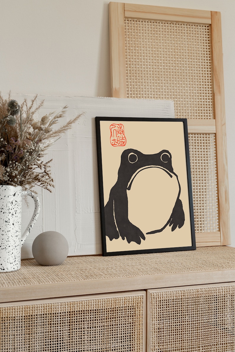 May include: Black and white illustration of a frog in a black frame on a light wood surface. The frog is on a cream background with a red stamp in the top left corner. The surface is a light wood with a woven pattern.