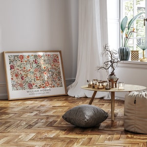 May include: A framed print of a floral pattern with the text "William Morris - Morris & Company, 1862 -" at the bottom. The print is hanging on a wall in a room with a wooden floor, a white curtain, and a small table with candles.