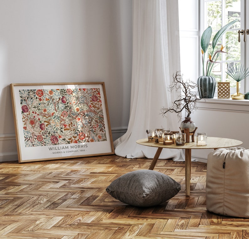 May include: A framed print of a floral pattern with the text "William Morris - Morris & Company, 1862 -" at the bottom. The print is hanging on a wall in a room with a wooden floor, a white curtain, and a small table with candles.