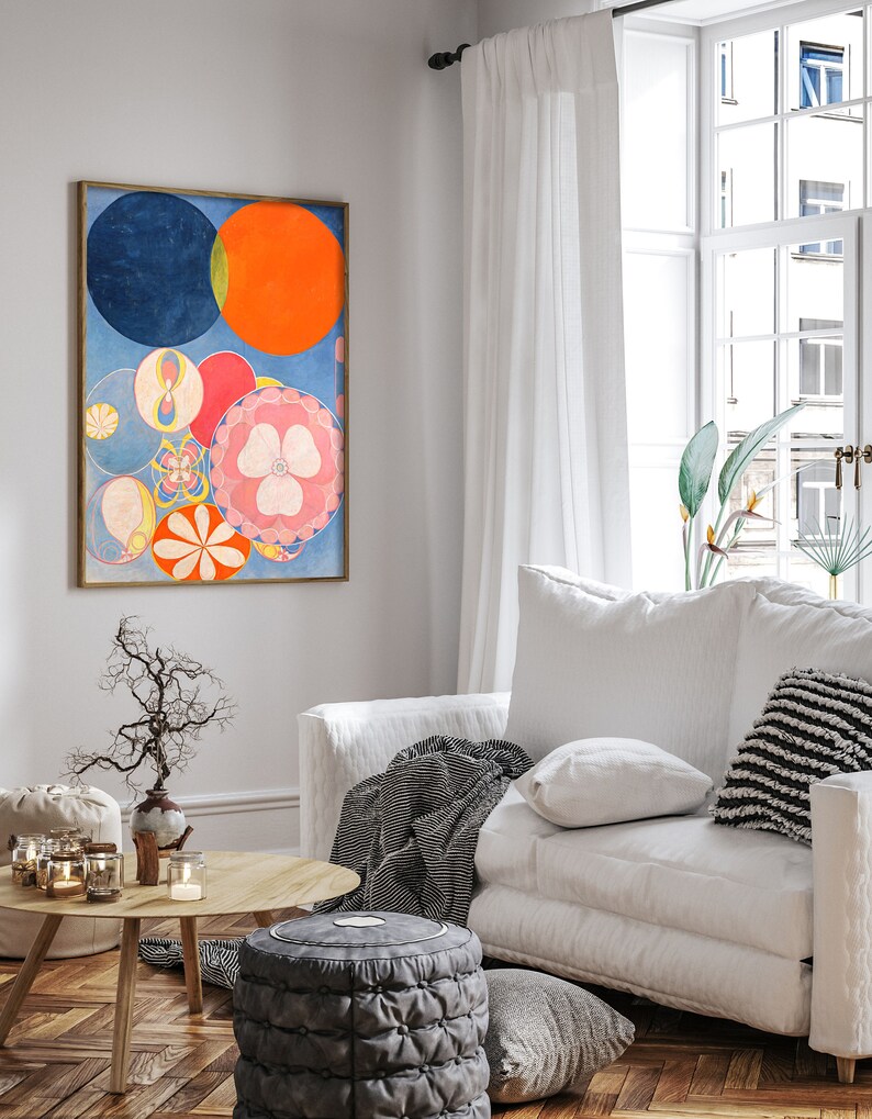 May include: An abstract art print in a gold frame features a composition of colorful circles and floral shapes. The artwork includes a large blue circle, an orange circle, and various smaller circles in pink, yellow, and red. The print hangs above a white sofa in a bright living room.