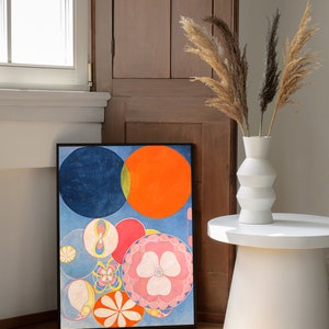 May include: A framed abstract art print featuring colorful circles in shades of blue, orange, red, and pink. The artwork is displayed on a wooden floor, leaning against a wooden door. A white vase with dried plants sits on a white side table.