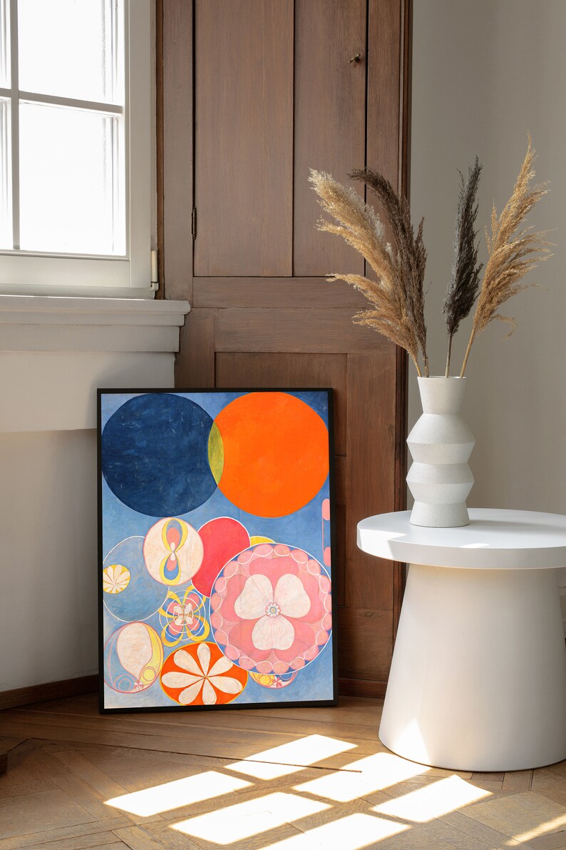 May include: A framed abstract art print featuring colorful circles in shades of blue, orange, red, and pink. The artwork is displayed on a wooden floor, leaning against a wooden door. A white vase with dried plants sits on a white side table.