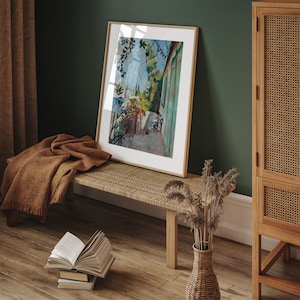 May include: A framed painting of a green building with a white door and a view of a garden. The painting is hanging on a green wall above a wooden bench with a woven seat. A vase with dried flowers sits on the floor next to the bench.