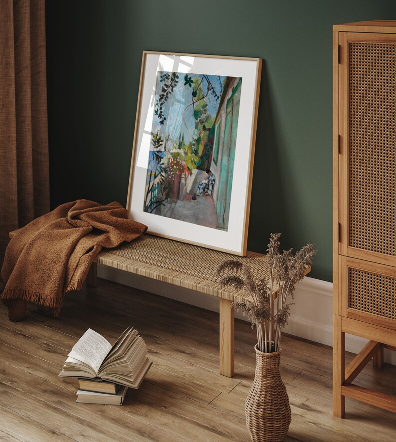 May include: A framed painting of a green building with a white door and a view of a garden. The painting is hanging on a green wall above a wooden bench with a woven seat. A vase with dried flowers sits on the floor next to the bench.