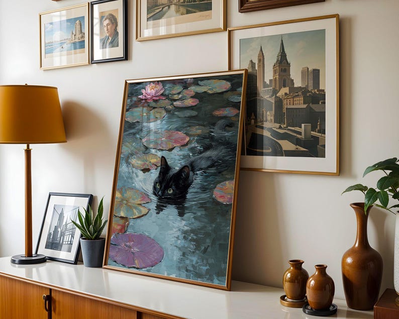 May include: A framed art print depicts a black cat swimming in a pond with lily pads and a pink water lily. The artwork is displayed on a white surface with other framed art, a lamp, and decorative vases.