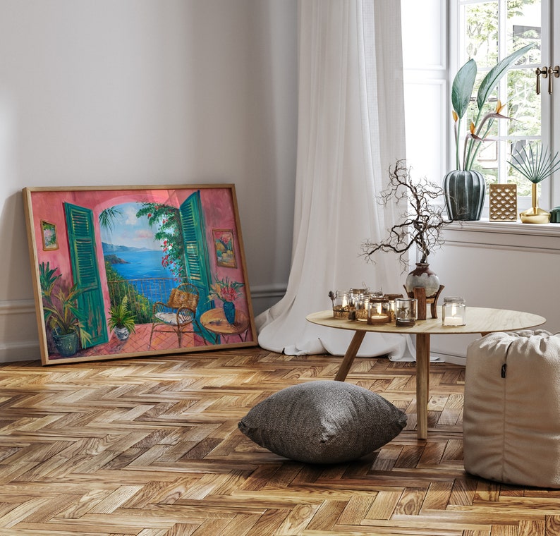 May include: A framed painting of a pink and green room with an open window overlooking a blue ocean. The painting is hanging on a white wall with a wooden floor. There is a small table with candles and a gray pillow on the floor.