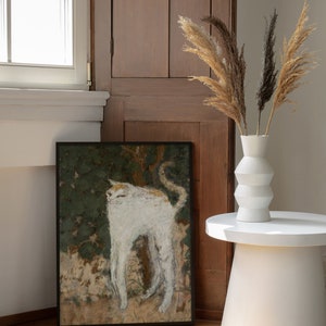 May include: A framed black and white painting of a white cat standing in a field of green foliage. The painting is hanging on a wall next to a wooden cabinet. A white vase with dried pampas grass is on a white side table.