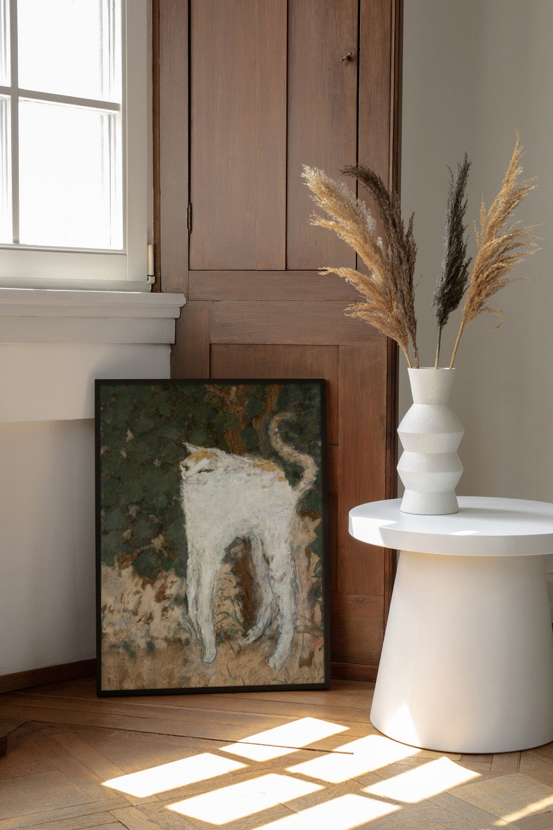 May include: A framed black and white painting of a white cat standing in a field of green foliage. The painting is hanging on a wall next to a wooden cabinet. A white vase with dried pampas grass is on a white side table.