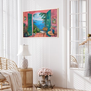 May include: A painting of a pink building with an open window overlooking a blue ocean. The window has green shutters and a wicker chair is sitting on the balcony. The painting is framed in gold.