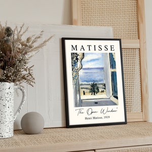 May include: A black framed print of a painting by Henri Matisse titled "The Open Window". The painting depicts a view from a window with a blue sky, sea, and palm tree.