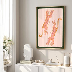 May include: A framed illustration of two pink leopards with orange spots jumping in the air against a light pink background. The frame is gold with a dark green mat.