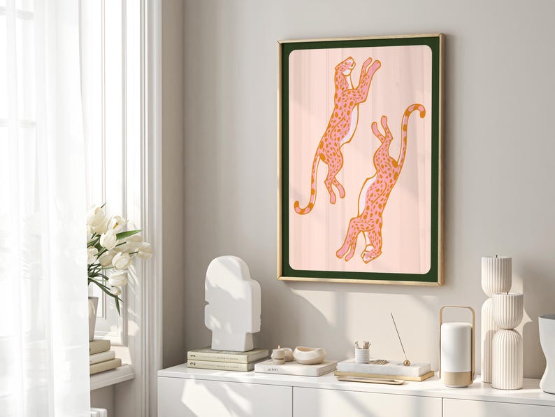 May include: A framed illustration of two pink leopards with orange spots jumping in the air against a light pink background. The frame is gold with a dark green mat.