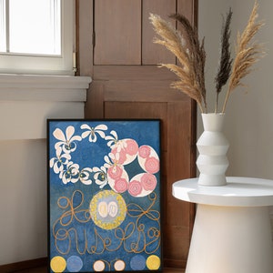 May include: A framed abstract painting with a blue background, white and pink flowers, and gold swirls. The painting is hanging on a wall next to a wooden cabinet and a white vase with dried pampas grass.