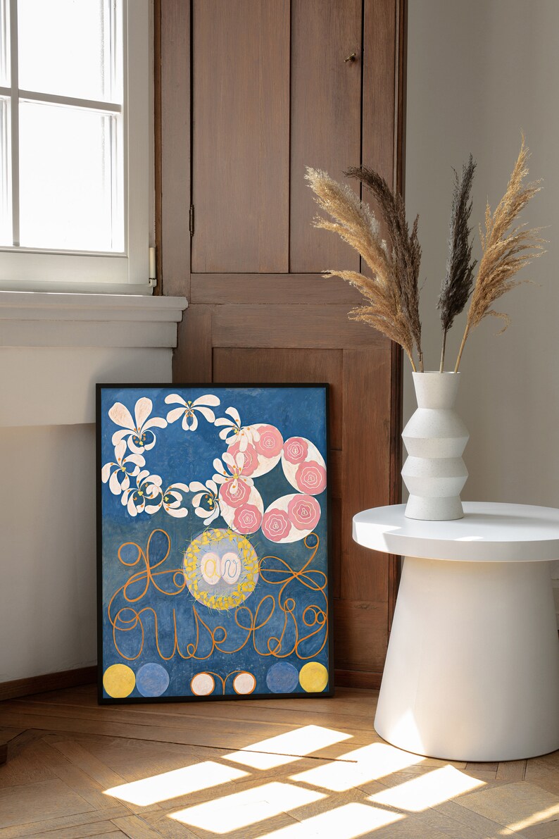 May include: A framed abstract painting with a blue background, white and pink flowers, and gold swirls. The painting is hanging on a wall next to a wooden cabinet and a white vase with dried pampas grass.