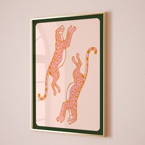 May include: A framed print featuring two pink and orange cheetahs with black spots, leaping in mid-air against a light pink background.