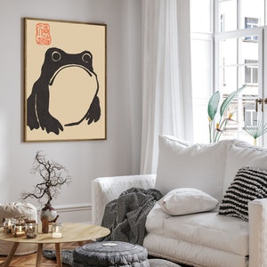 May include: A black and white illustration of a frog with a beige background. The image is framed in a light wood frame and hanging on a white wall. The image is in a living room setting with a white couch, a coffee table, and a plant.