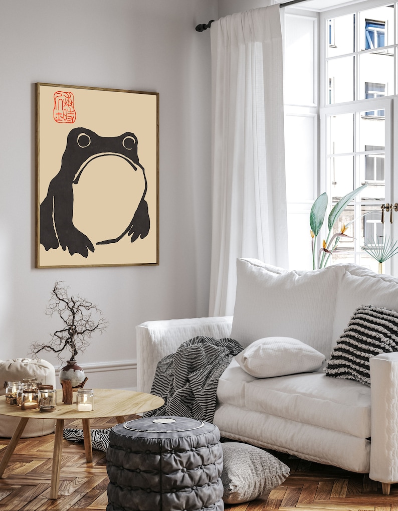 May include: A black and white illustration of a frog with a beige background. The image is framed in a light wood frame and hanging on a white wall. The image is in a living room setting with a white couch, a coffee table, and a plant.