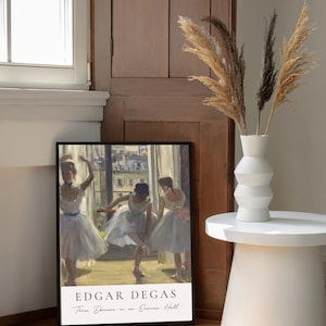 May include: A framed print of a painting by Edgar Degas titled "Three Dancers in an Exercise Hall". The painting depicts three ballerinas in white tutus practicing in a dance studio. The print is framed in black and has a white mat.