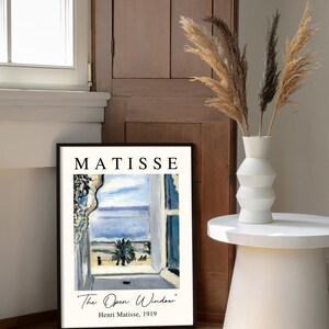 May include: A black framed print of a painting by Henri Matisse titled "The Open Window". The painting depicts a view from a window overlooking a blue sea and a palm tree.